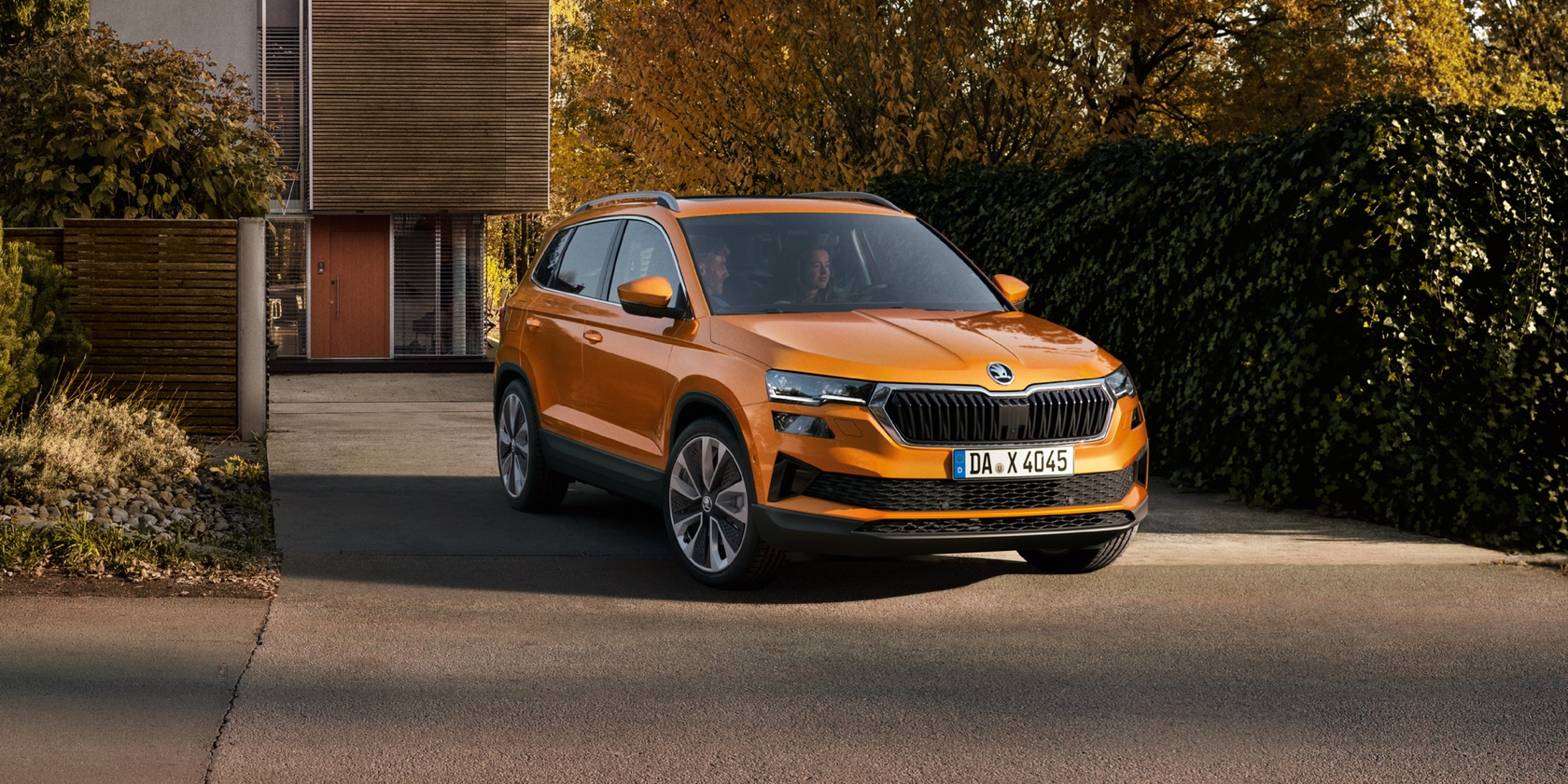 Orangefarbener Škoda Karoq in seitlicher Frontansicht steht in einer Einfahrt vor einem modernen Wohnhaus. Ein Mann sitzt auf dem Beifahrersitz, eine Frau am Steuer.