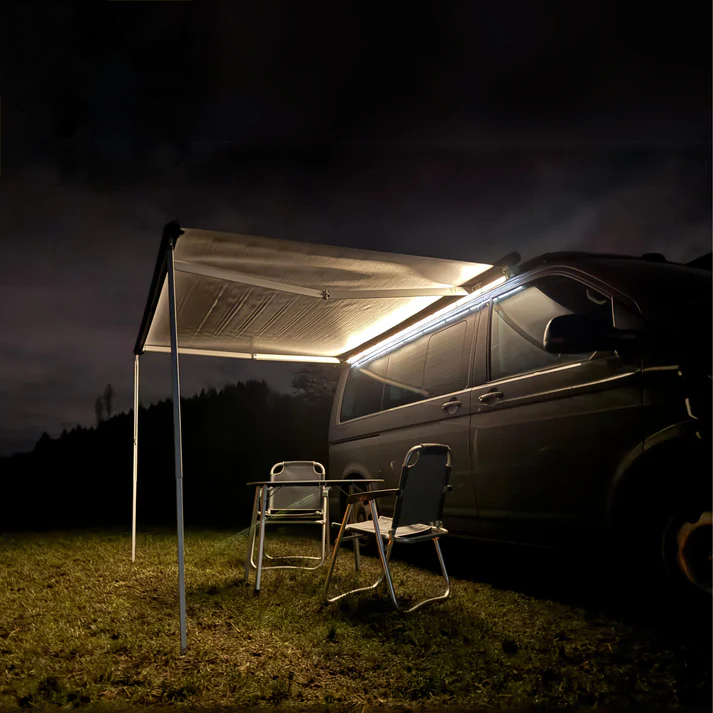Dunkelgrauer VW Ocean in seitlicher Frontansicht bei Nacht. Ausgefahrene Markise mit Markisenbeleuchtung von CALIFAKTUR. Vor dem Camper stehen zwei Campingstühle und ein kleiner Campingtisch auf dem Rasen.