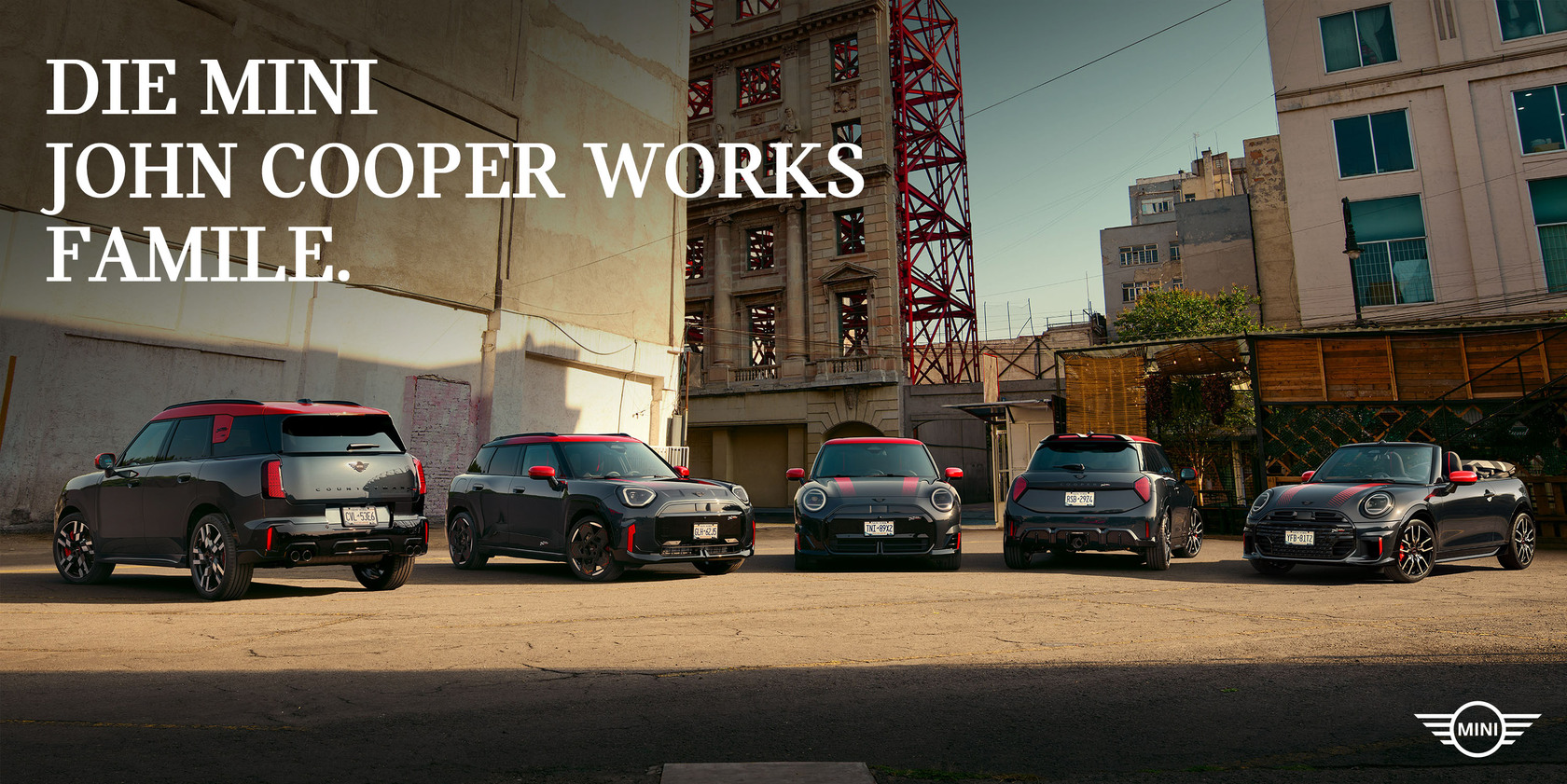 ‚Die MINI John Cooper Works Familie‘ mit fünf sportlichen MINI John Cooper Works Modellen nebeneinander in urbaner Industrieumgebung, schwarz-rote Designakzente im Vordergrund.