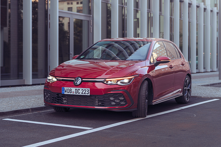 Roter VW Golf GTI steht auf einem Parkplatz vor einem modernen Bürogebäude. Frontansicht mit charakteristischem GTI-Design und sportlichen Details.
