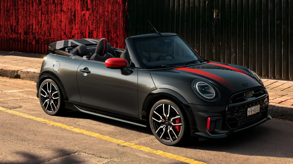 Schwarzer MINI John Cooper Works Cabrio in Seiten- und Frontansicht mit roten Folierungen und Akzenten, geparkt vor einem roten Gebäude. Das Dach des Cabrios ist geöffnet.