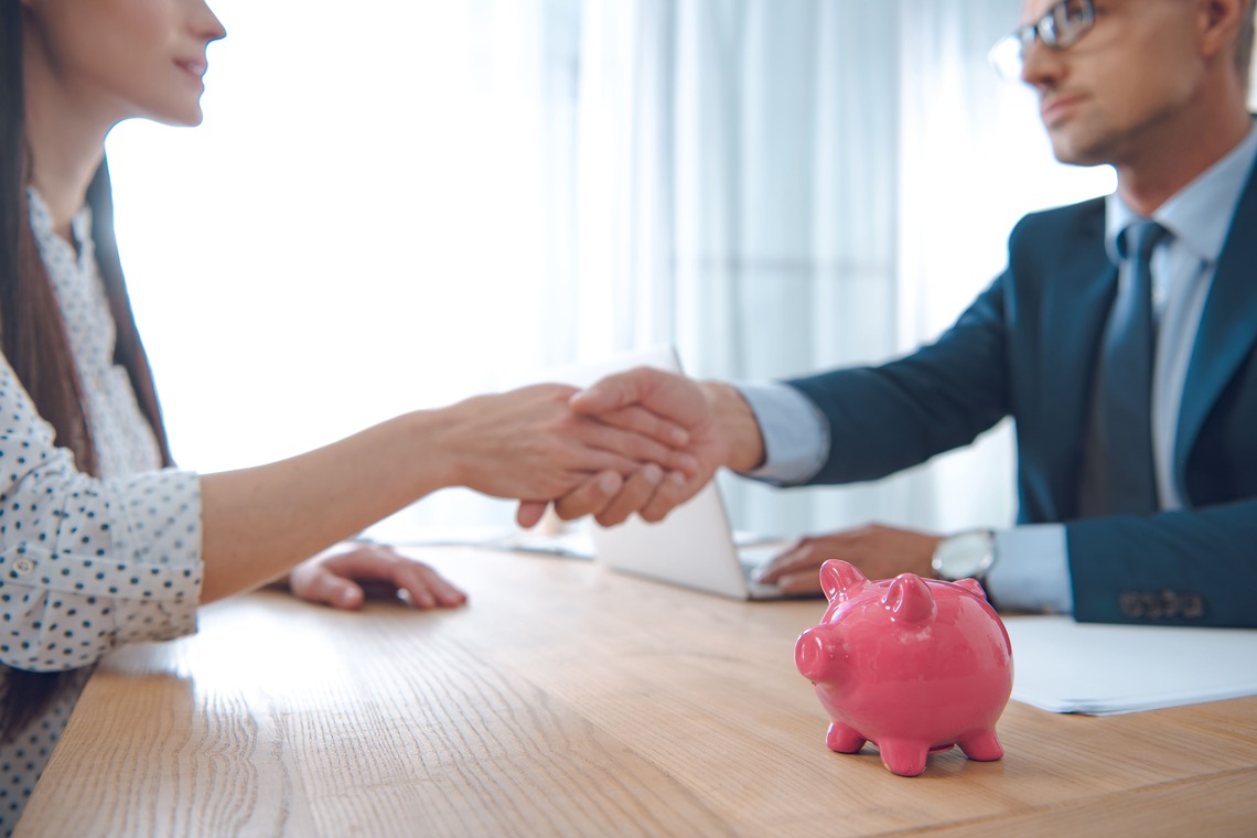 Geschäftliches Händeschütteln zwischen Kundin und Berater – im Vordergrund ein pinkes Sparschwein als Symbol für Finanzberatung.
