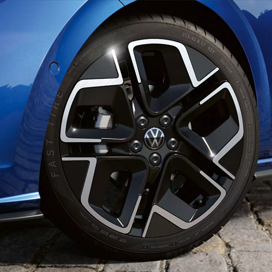 Detailaufnahme einer schwarz-silbernen VW-Leichtmetallfelge mit markantem Zierschliff und VW-Logo auf einem blauen Fahrzeug.
