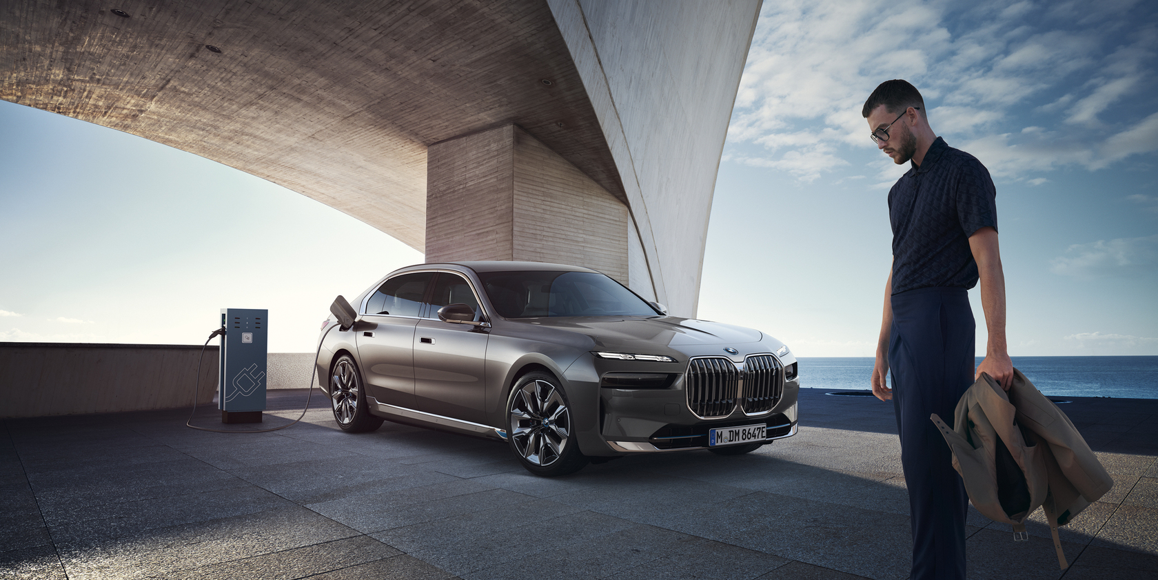 BMW i7 an einer Ladesäule angeschlossen, unter einem modernen Gebäude. Rechts im Bild steht ein Mann mit Brille und einer braunen Jacke in der Hand. Im Hintergrund ist das Meer zu sehen.