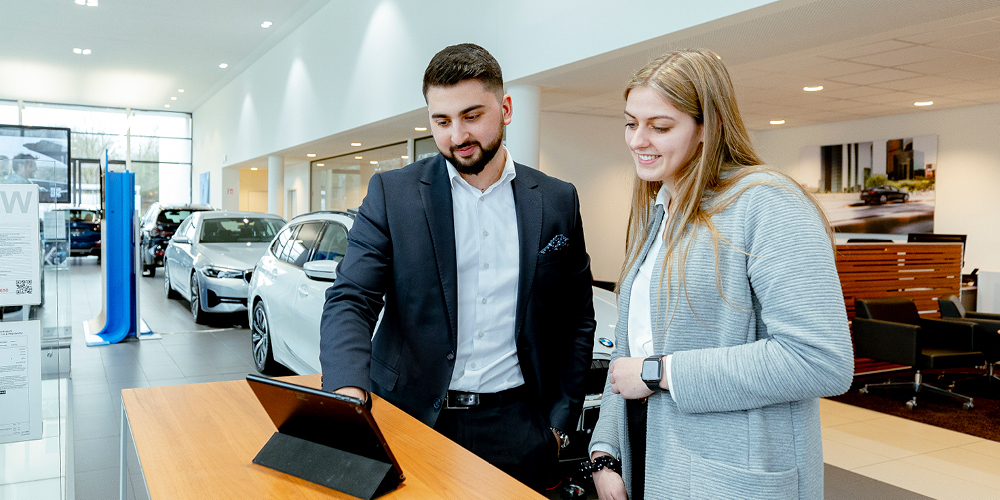 Zwei Personen stehen in einer BMW-Verkaufshalle und schauen gemeinsam auf ein Tablet. Im Hintergrund verschiedene BMW-Modelle