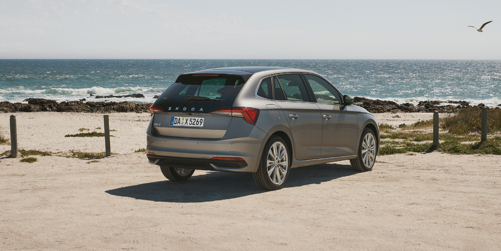 Ein grauer Škoda Scala in Heck- und Seitenansicht steht auf einem sandigen Parkplatz mit Blick auf das Meer.