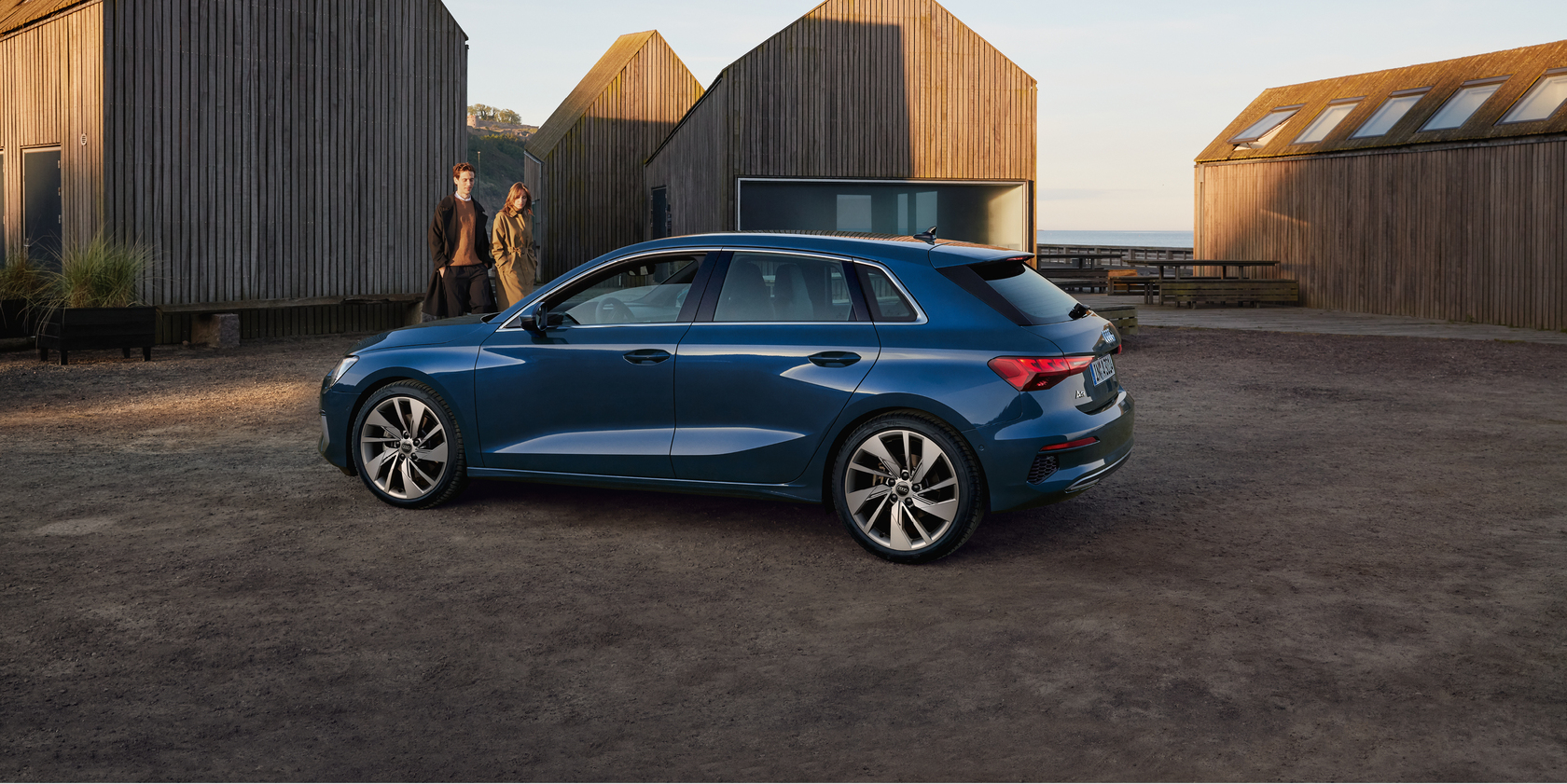 Blauer Audi A3 Sportback in Seitenansicht, geparkt vor modernem Haus. Im Hintergrund gehen ein Mann und eine Frau in Richtung Auto