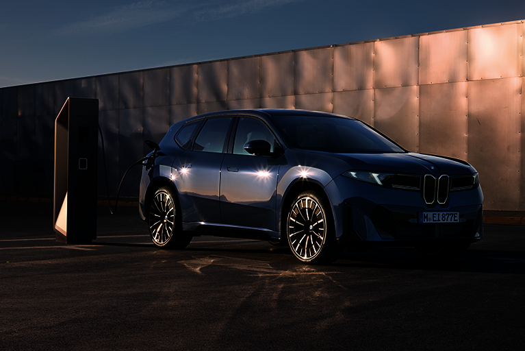 Der neue vollelektrische BMW iX3 in dunklem Blau steht an einer modernen Ladesäule und wird geladen – urbaner Hintergrund mit metallischer Wand bei Sonnenuntergang.