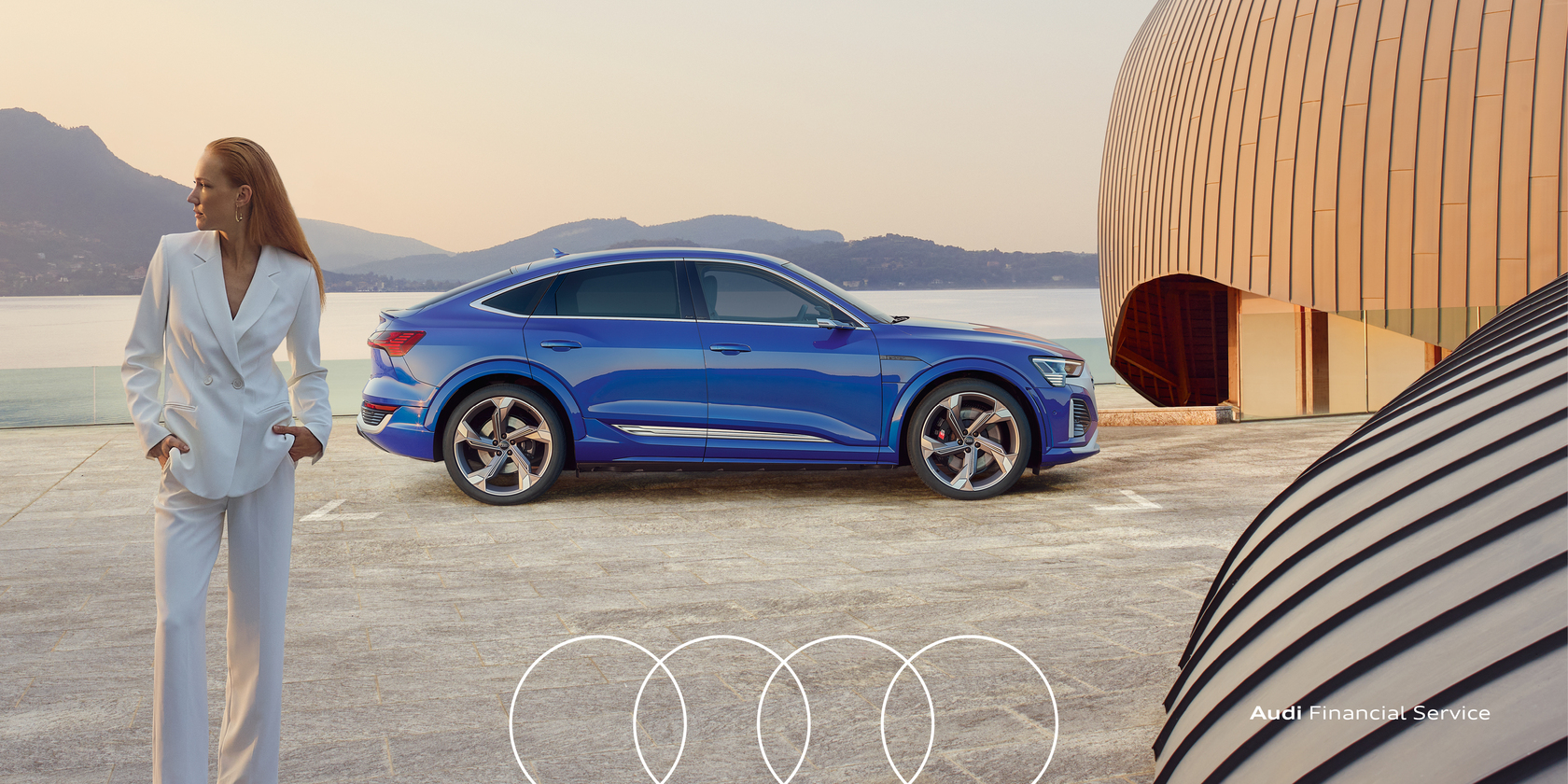 Blauer Audi e-tron Sportback in Seitenansicht vor moderner Architektur und einem See, Frau in weißem Anzug steht im Vordergrund. Am unteren Rand des Bildes die Audiringe sowie der weiße Schriftzug "Audi Financial Service"
