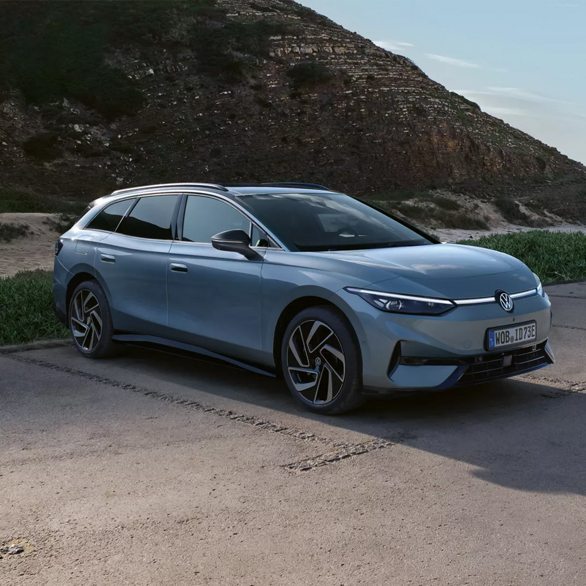 Hellblauer Volkswagen ID.7 Tourer in Seiten- und Frontansicht, geparkt auf einem Parkplatz vor einer Felswand. 