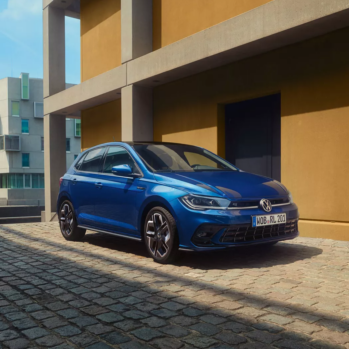 Blauer VW Polo R-Line in seitlicher Frontansicht geparkt auf Kopfsteinpflaster vor orangener Hauswand  bei Sonnenschein.
