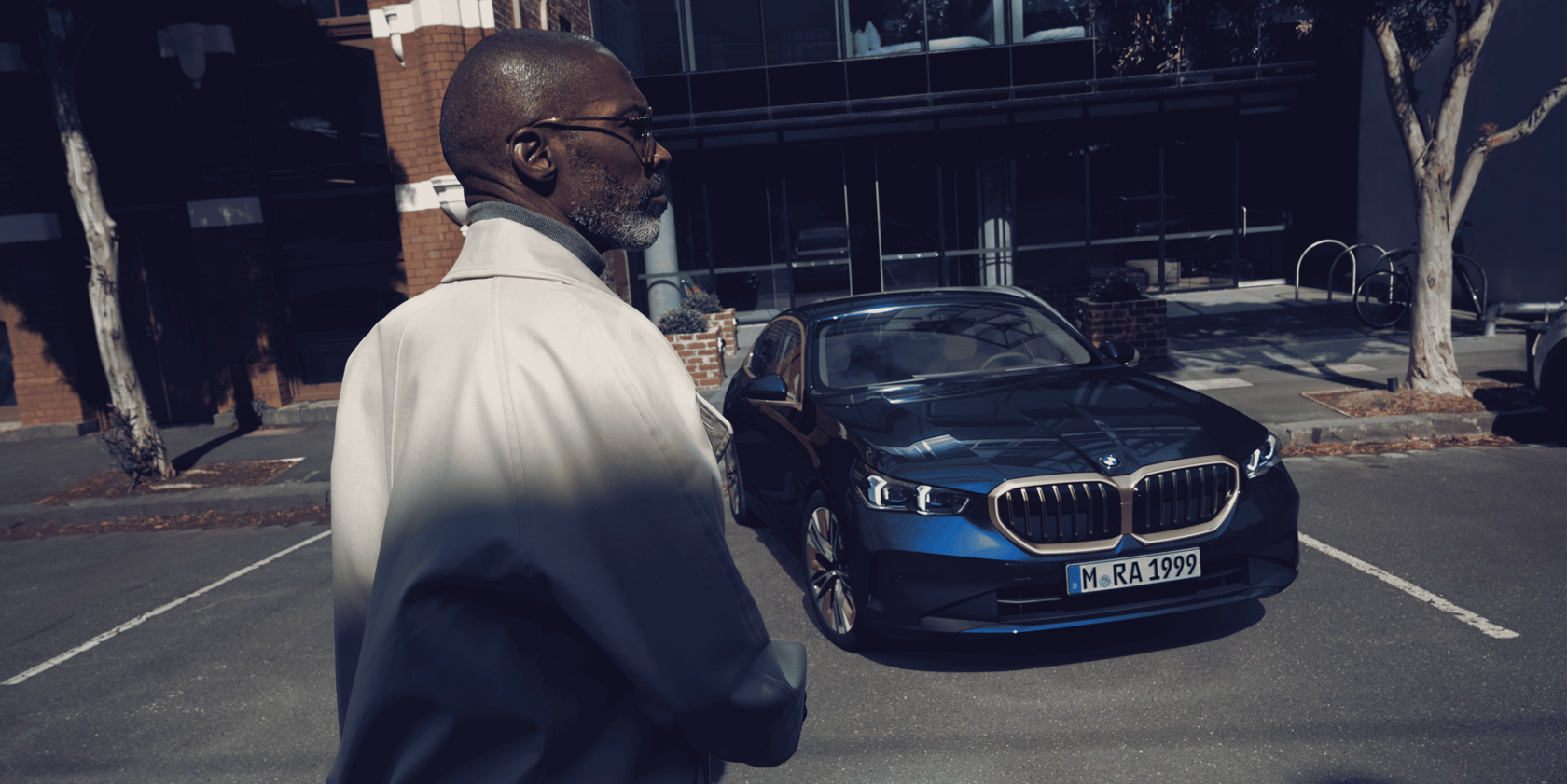 Mann im Vordergrund, seitlich abgewandt, im Hintergrund ein blauer BMW 5er in Frontansicht auf einem Parkplatz vor einem modernen Gebäude.