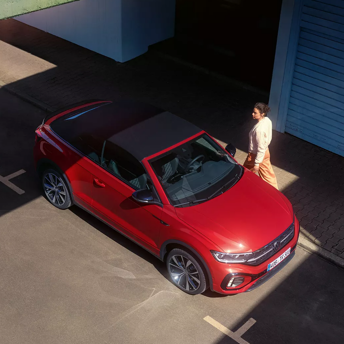 Rotes VW T-Roc Cabriolet mit geschlossenem Verdeck aus der Vogelperspektive, eine Frau geht auf dem Bürgersteig am geparkten Auto vorbei.