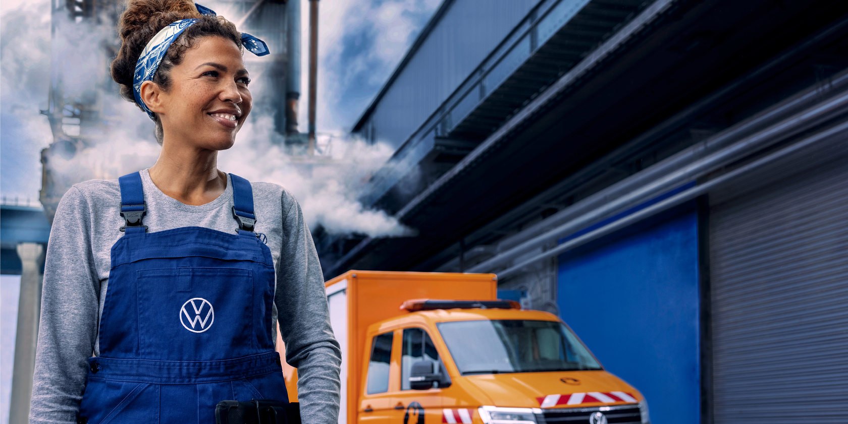 Lächelnde Frau in blauer Latzhose mit weißem VW Logo. Im Hintergund ein orangener VW Crafter Pritsche mit Aufbau und ein großes Industriegebäude aus dem Dampf aufsteigt
