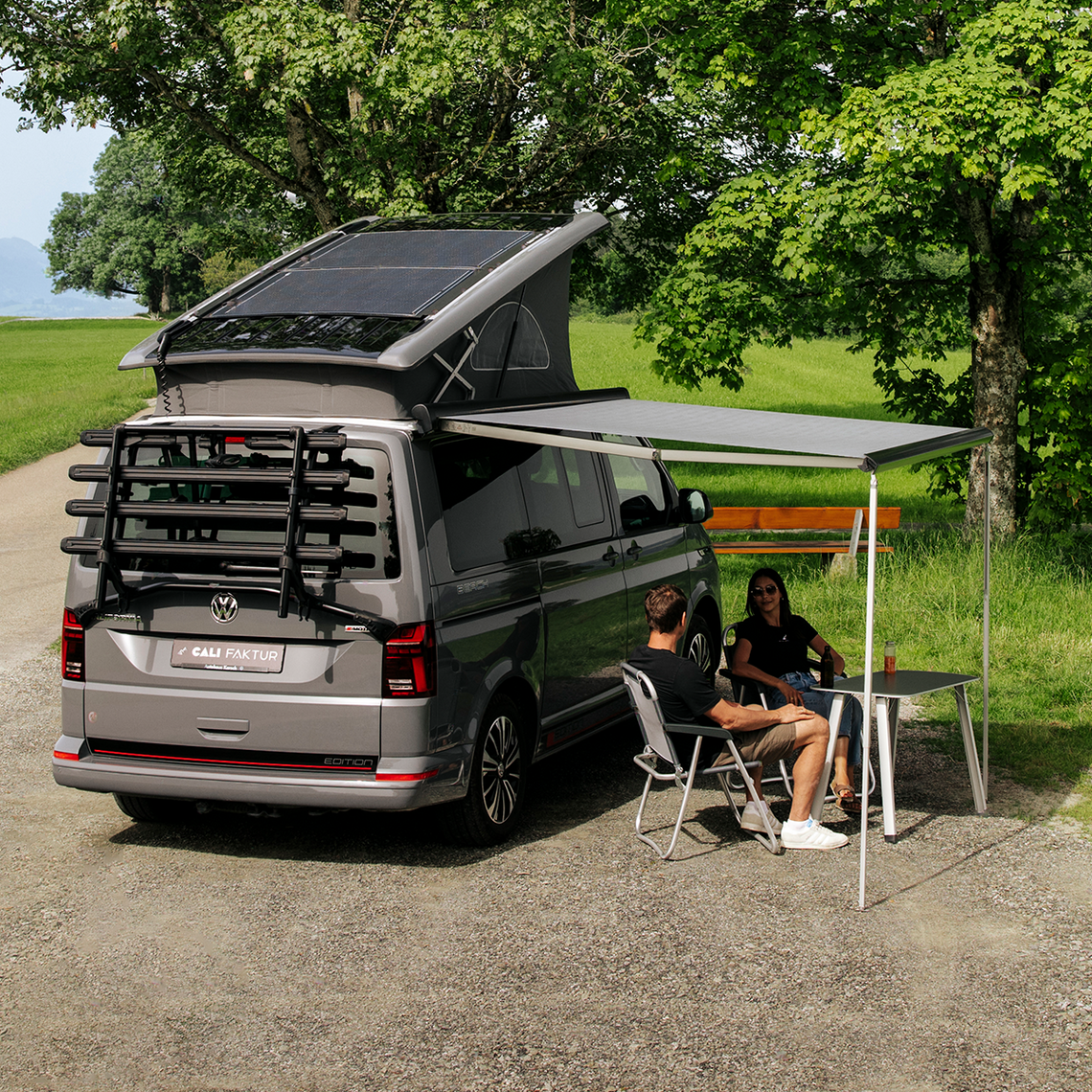 Grauer VW California Ocean in Heckansicht. Ausgeklapptes Aufstelldach mit eingebauten Solarpanelen von CALIFAKTUR. Zwei Personen sitzen unter der ausgefahrenen Markise. Im Hintergrund eine grüne Wiese und Bäume.