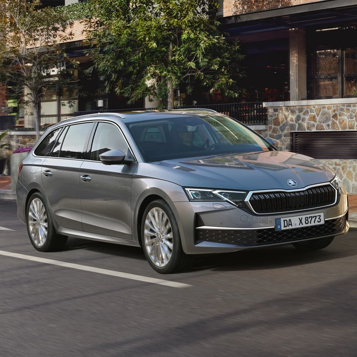Grauer Škoda Octavia Kombi fährt auf der Straße an einer modernen Steinfassade vorbei.