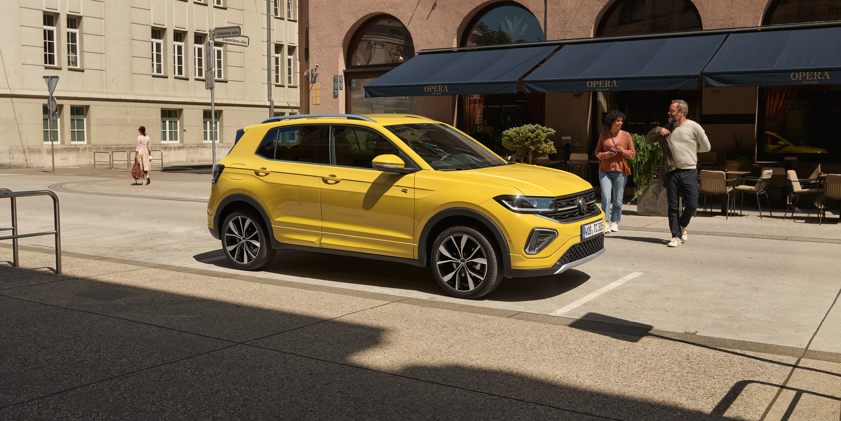 Gelber Volkswagen T-Cross in Seitenansicht geparkt am Straßenrand vor einem Café mit blauen Markisen, zwei Personen gehen vor dem Auto über die Straße.