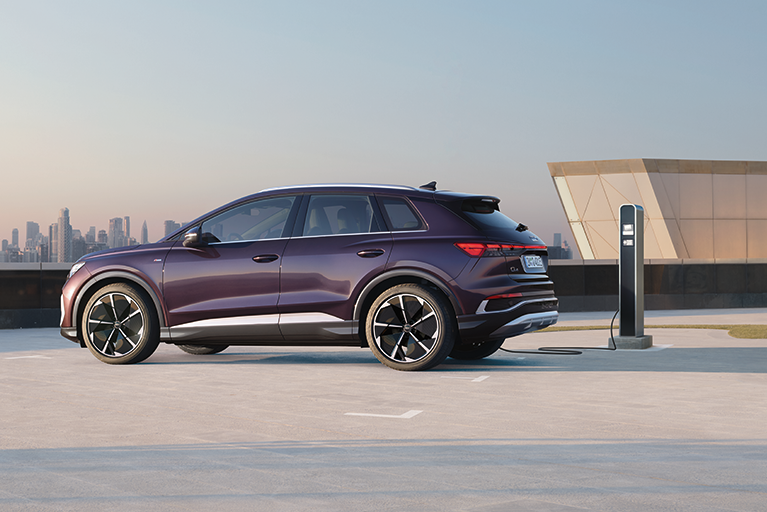 Lila Audi Q4 e-tron SUV steht seitlich an einer Ladesäule auf einem offenen Parkdeck mit Blick auf eine moderne Skyline im Hintergrund.
