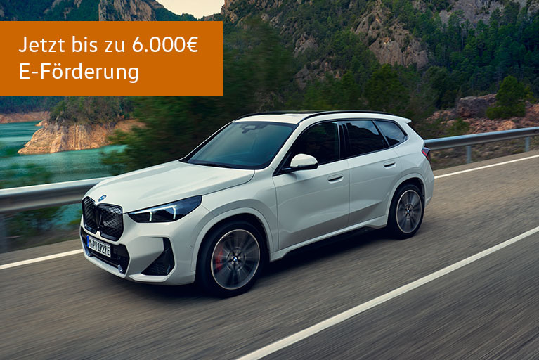 Weißer BMW iX1 Elektro-SUV fährt auf einer kurvigen Straße entlang eines Sees in Berglandschaft, mit Hinweis auf bis zu 6.000 Euro E-Förderung.