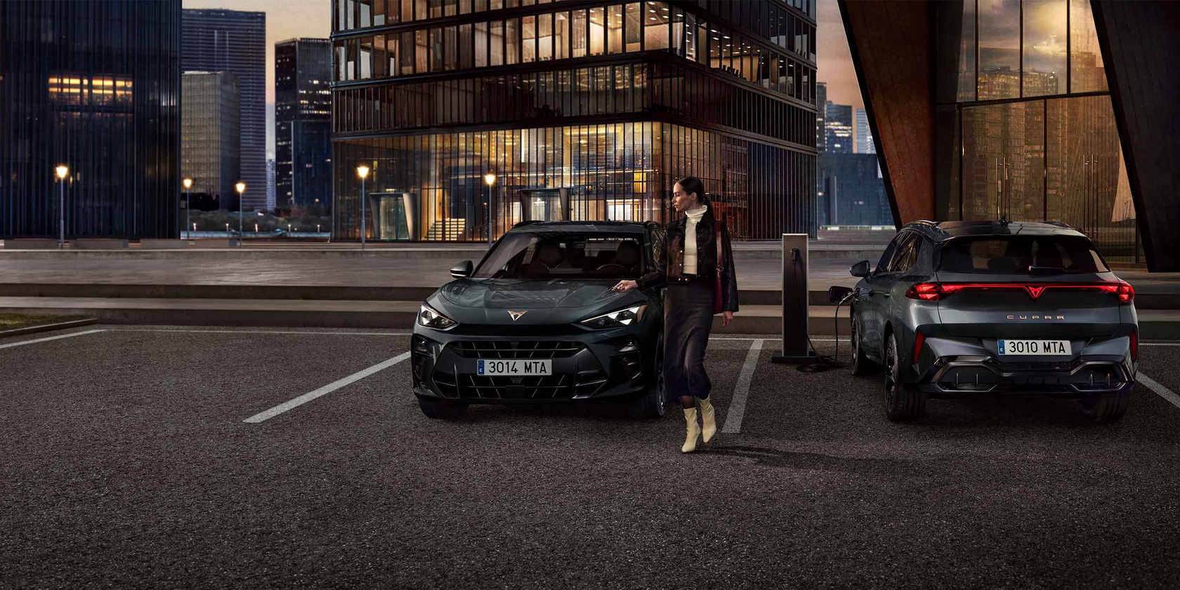 Zwei CUPRA Fahrzeuge stehen auf einem Parkplatz vor moderner Stadtkulisse; eines der Fahrzeuge ist an eine Ladesäule angeschlossen, während eine Frau zwischen den Autos steht.“