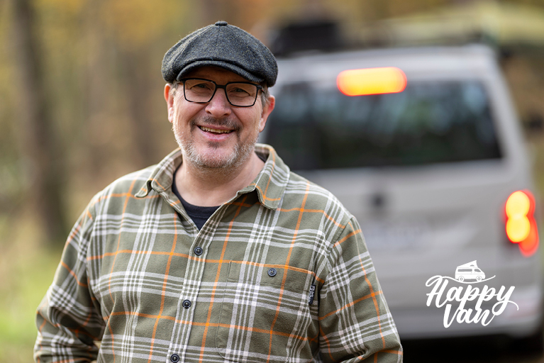 Ein lächelnder Mann mit Brille, Schiebermütze und kariertem Hemd (Wölle). Im unscharfen Hintergrund ist ein weißer Van mit eingeschaltetem Bremslicht zu sehen. Rechts unten im Bild das Happy Van Logo