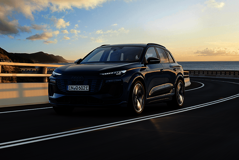 Dunkelblauer Audi Q6 e-tron fährt auf kurviger Küstenstraße mit Blick aufs Meer bei Sonnenuntergang.