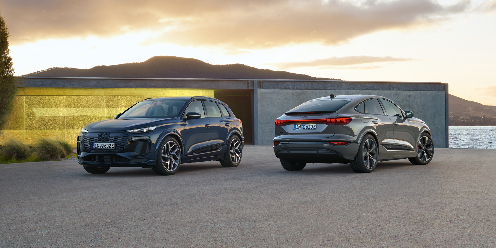 ein dunkelblauer Audi Q6 e-tron in Front- und Seitenansicht und ein silberfarbener Audi Q6 e-tron Sportback in Heck- und Seitenansicht vor malerischer Kulisse