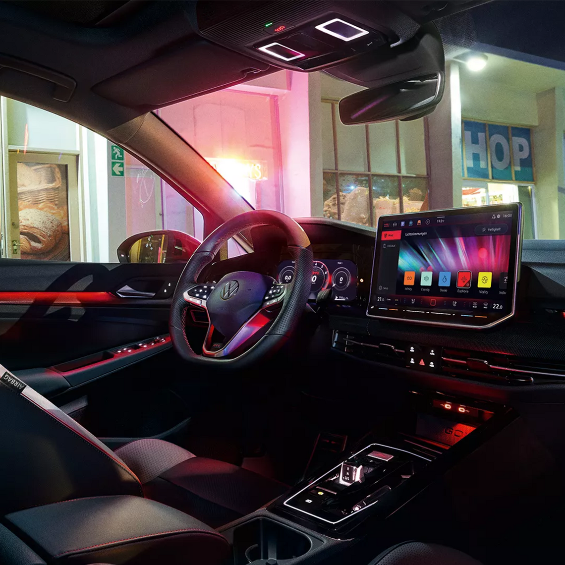 Cockpit eines VW Golf GTI bei Nacht mit sportlichem Lenkrad, großem Touchscreen-Display mit farbigem Menü, roter Ambientebeleuchtung und Blick nach draußen auf ein beleuchtetes Schild eines Geschäftes