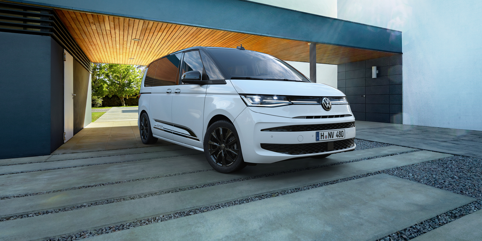 Weißer Volkswagen Multivan Edition T7 in seitlicher Frontansicht, geparkt vor der Eingangstür eines modernen Gebäudes.