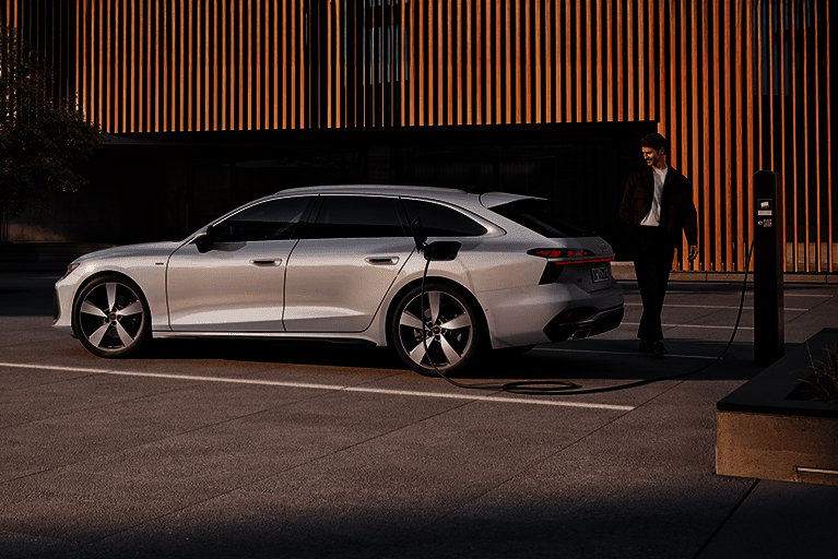Seitenansicht eines weißen Audi A6 Avant e-hybrid, angeschlossen an eine Ladesäule. Ein Mann steht daneben.
