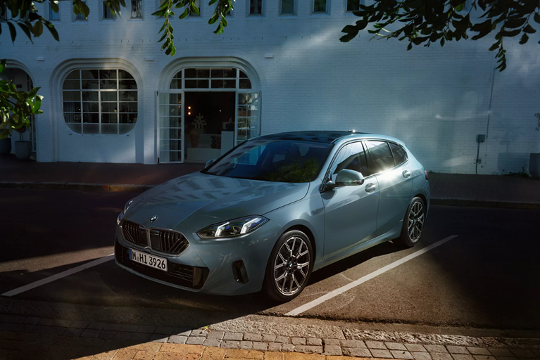 Grün grauer BMW 1er steht auf einem Parkplatz vor einem weißen Gebäude mit großen Rundbogenfenstern. Im sportlichem Design und Bicolor Leichtmetallfelgen.