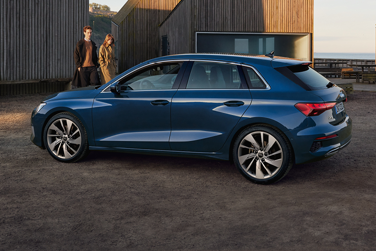 Blauer Audi A3 Sportback in Seitenansicht, geparkt vor einem modernen Haus. Im Hintergrund gehen ein Mann und eine Frau in Richtung Auto