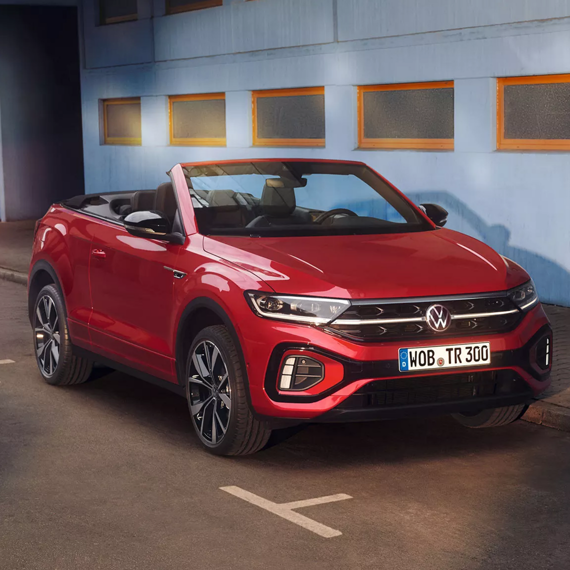 Rotes VW T-Roc Cabriolet mit geöffnetem Verdeck in Front- und Seitenansicht, geparkt vor einer hellblauen Hauswand mit kleinen Fenstern.
