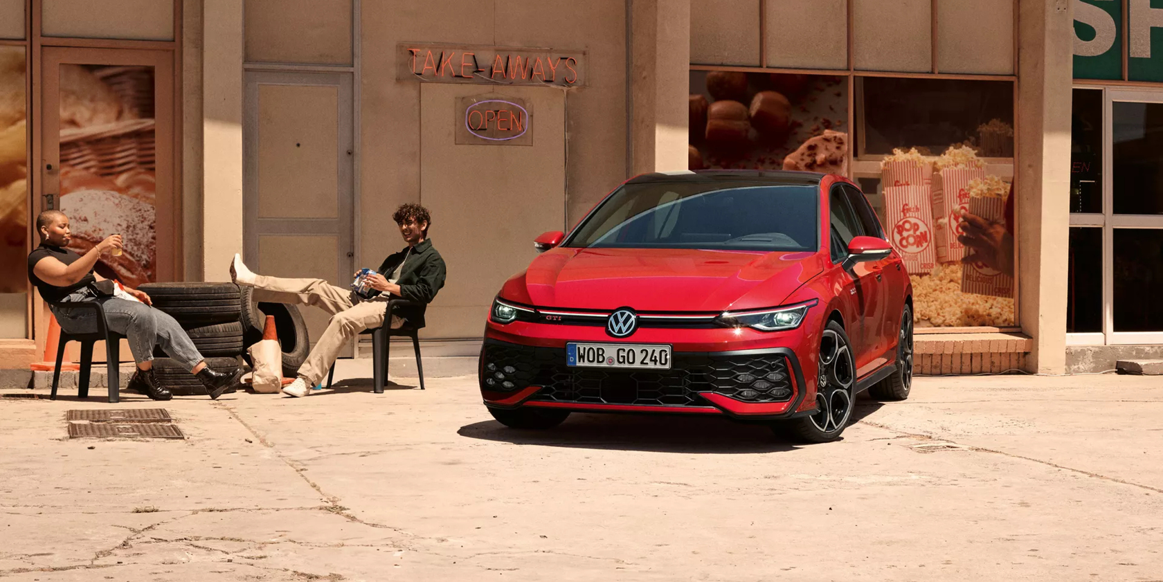 Roter Volkswagen Golf GTI in Frontansicht vor einem Imbiss mit "Take-Aways"-Schild, zwei Personen  sitzen neben dem Auto und trinken etwas.