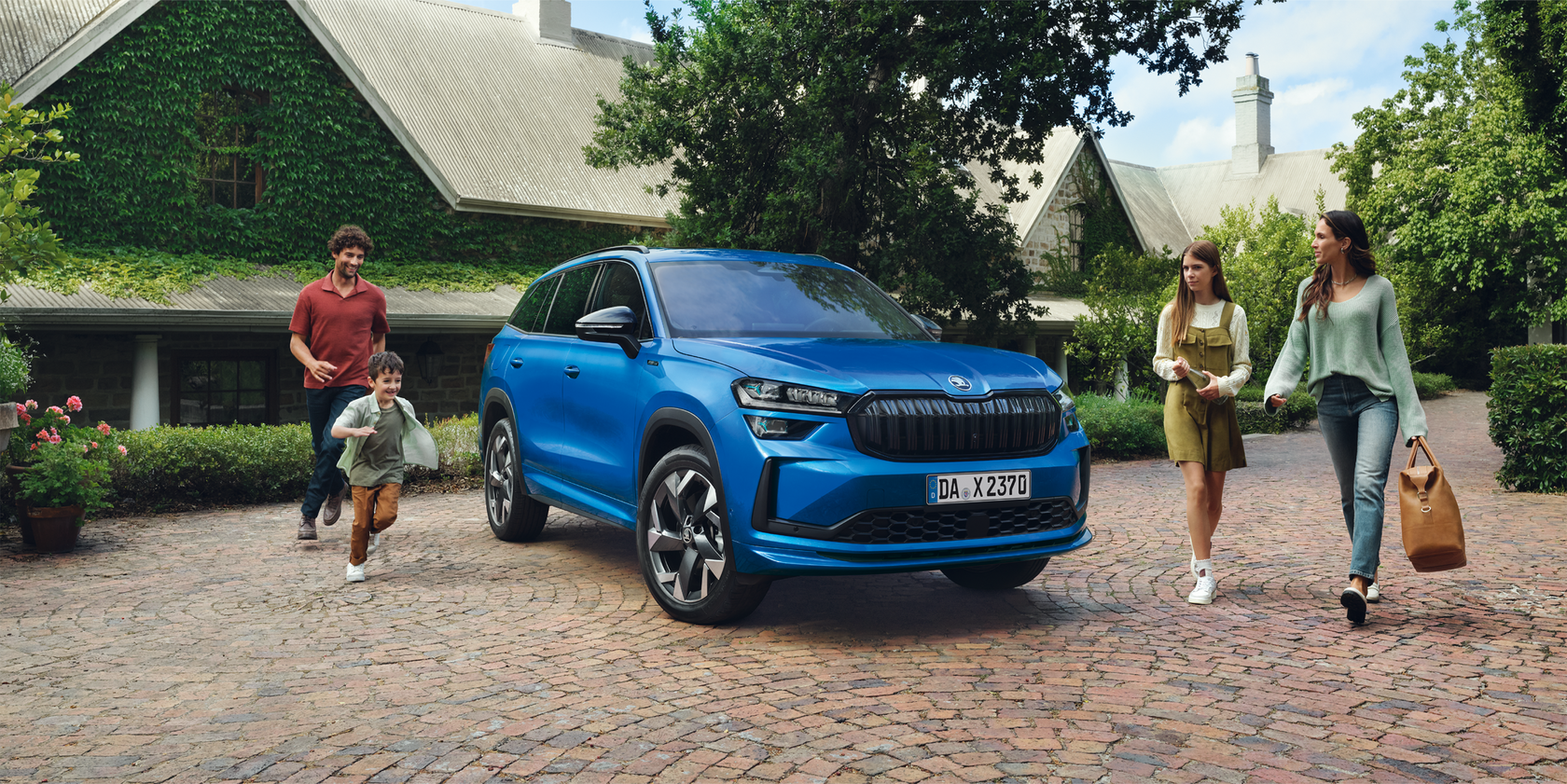 Blauer Skoda Kodiaq geparkt vor einem großen Wohnhaus, eine vierköpfige Familie läuft um das Autos herum