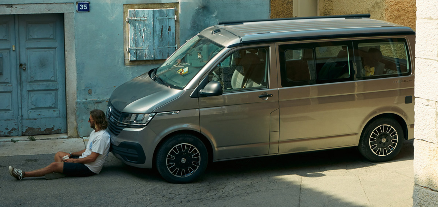 Grauer VW California Beach, geparkt am Straßenrand vor einem hellblauen Haus mit Holztür und Holzfenster, ein Mann sitzt auf dem Boden vor dem Auto, an die Front des Campers gelehnt.