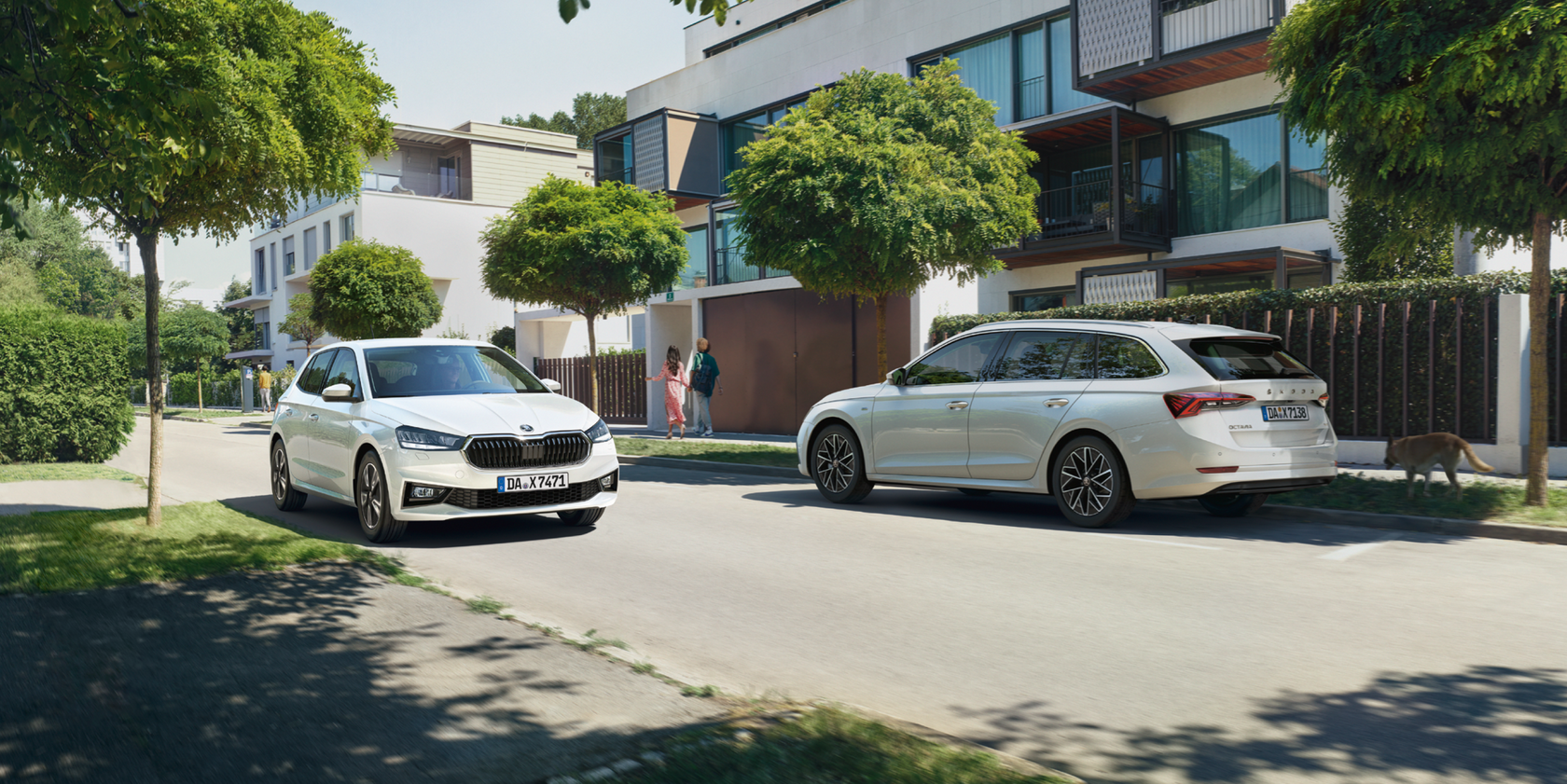 Zwei Škoda Modelle fahren auf einer Straße in einem begrünten Wohngebiet in entgegengesetzte Richtungen. Ein weißer Škoda Fabia in Frontansicht und ein weißer Škoda Octavia Kombi in Seiten- und Heckansicht. Im Hintergrund läuft ein Paar sowie ein Hund auf dem Bürgersteig.