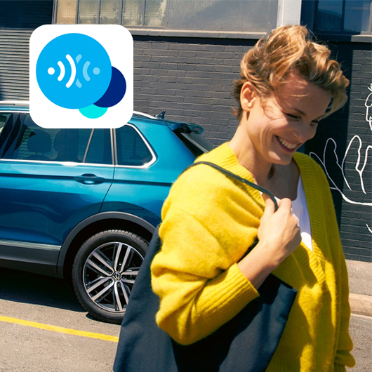 Eine Frau mit gelber Jacke und dunkler Tasche geht lächelnd an einem blauen Auto vorbei, das vor einer dunklen Hauswand steht. Oben links im Bild ist ein blaues App-Symbol mit Kreisen zu sehen. 