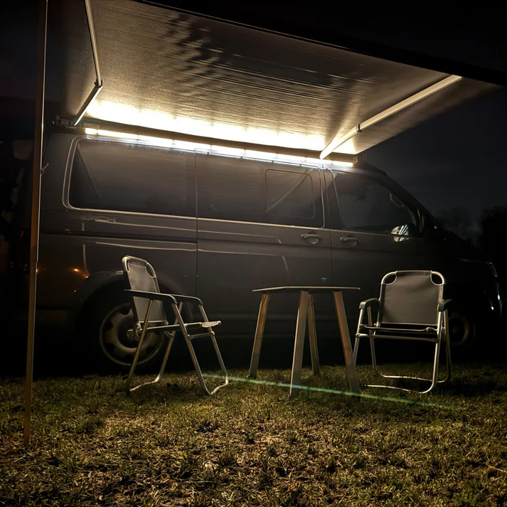 Dunkelgrauer VW California Ocean in Seitenansicht bei Nacht. Ausgefahrene Markise mit Markisenbeleuchtung von CALIFAKTUR. Vor dem Camper stehen zwei Campingstühle und ein kleiner Campingtisch auf dem Rasen.