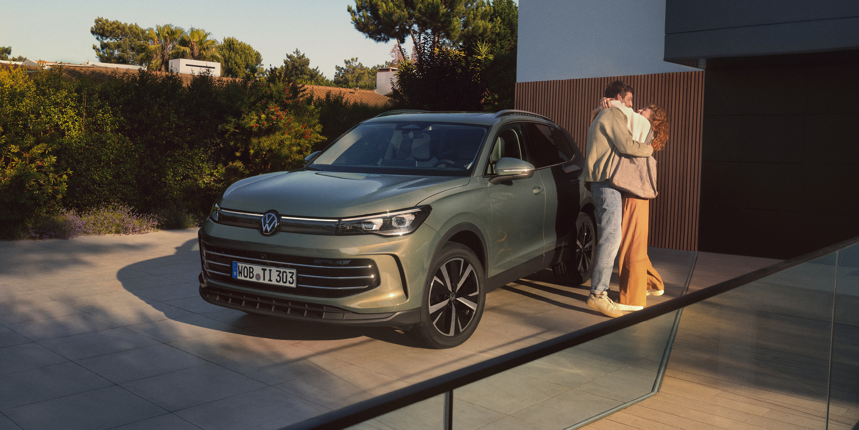 Grauer Volkswagen Tiguan in Front- und Seitenansicht geparkt vor modernem Haus, ein Paar umarmt sich neben dem Auto