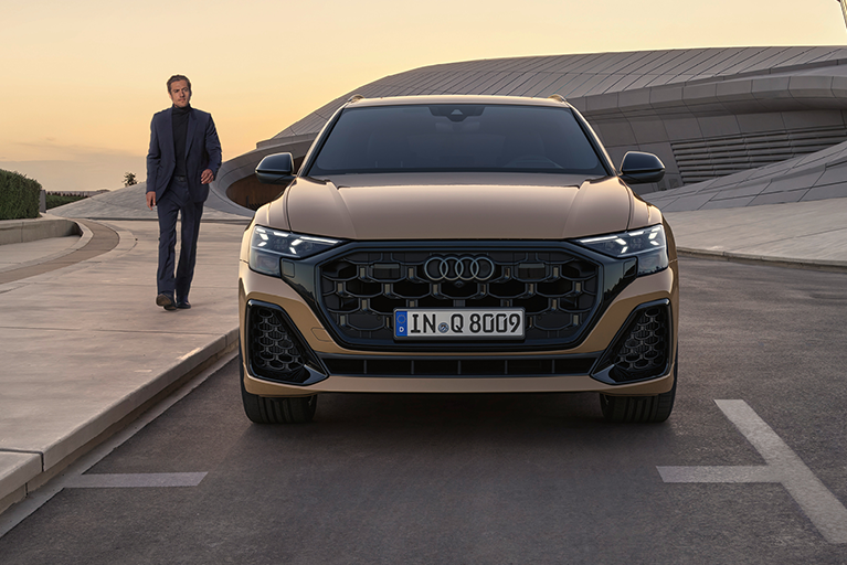 Braun/goldener Audi Q8 bei Sonnenuntergang in Frontalansicht, geparkt vor moderner Architektur. Rechts neben dem Auto ein Mann gehend auf dem Bürgersteig