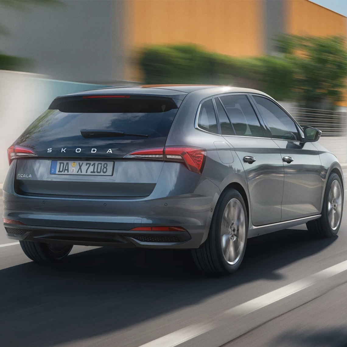 Ein grauer Škoda Scala in Heckansicht fährt auf einer Straße entlang einer modernen Gebäudefassade.