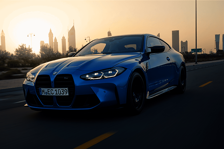 Dynamischer BMW M4 in blauer Lackierung fährt bei Sonnenuntergang durch eine moderne Stadtlandschaft.