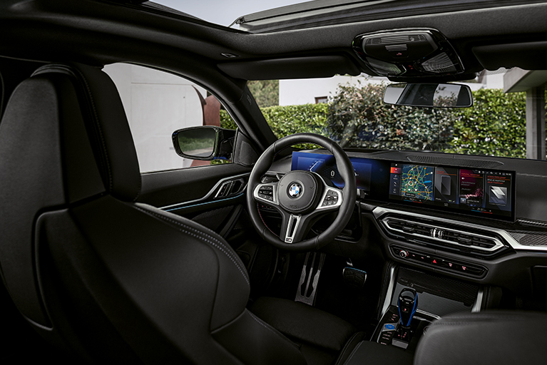 Innenraum eines BMW i4 M50 mit Curved Display, Multifunktionslenkrad und blauen Applikationen am Schalthebel. Perspektive von den Rücksitzen aus.