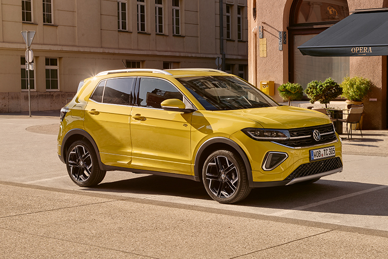 Gelber Volkswagen T-Cross schräg von vorn auf einem Stadtplatz vor Gebäuden und einem Café geparkt.