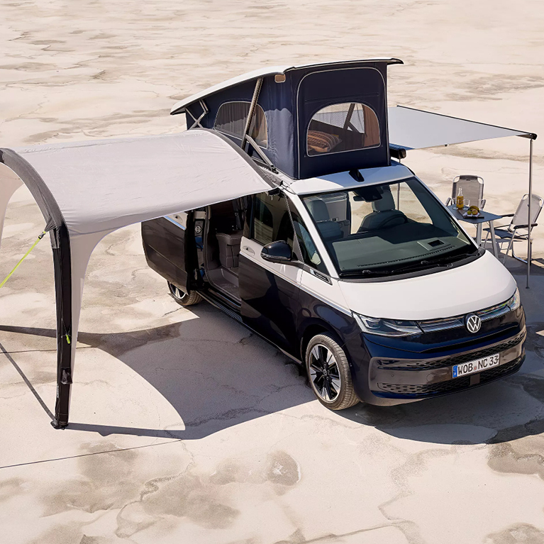 Schwarz-weißer VW California Beach mit ausgeklapptem Aufstelldach, zwei geöffneten Schiebetüren, einem Vorzelt auf der einen Seite sowie einer ausgefahrenen Markise auf der anderen Seite. Unter der Markise stehen zwei Campingstühle und ein Tisch.