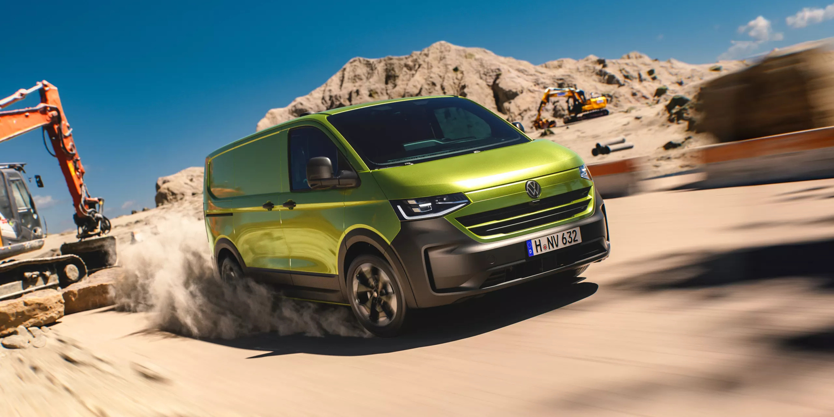 Grüner VW Transporter in seitlicher Frontansicht fahrend auf einer sandigen Baustelle, Bagger und Sandhügel im Hintergrund.