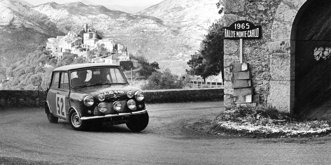 Historisches Schwarz-Weiß-Foto eines MINI Cooper S mit der Startnummer 52 bei der Rallye Monte Carlo 1965, der eine enge Kurve auf einer Bergstraße fährt – im Hintergrund ein Dorf in den französischen Alpen.
