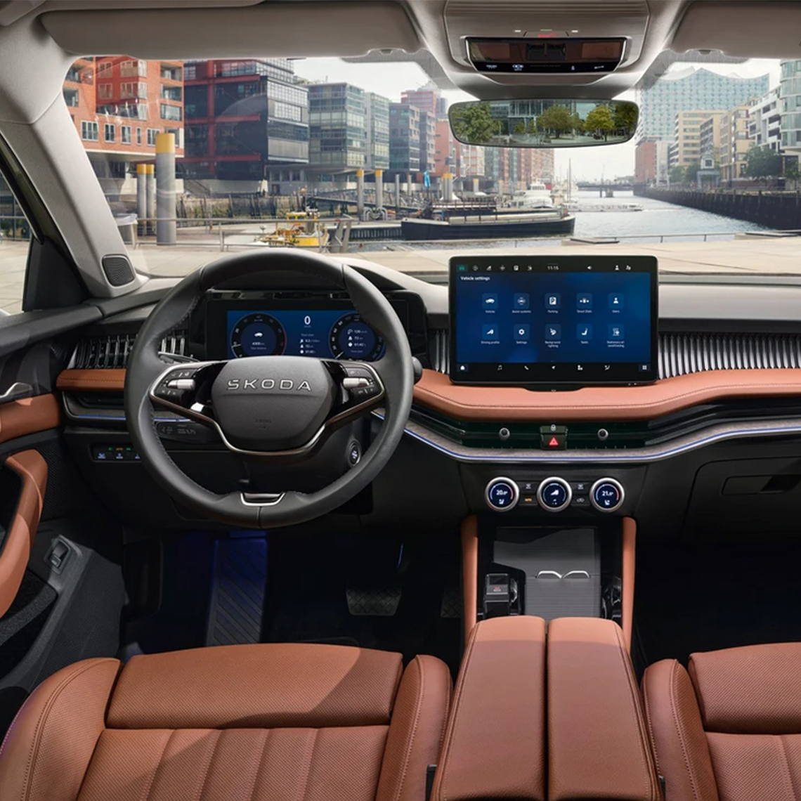 Innenraum eines Škoda-Fahrzeugs mit moderner, hochwertiger Ausstattung. Multifunktionslenkrad mit Škoda-Logo. Interieur in edlem Dunkelrot-Braun mit schwarzen Kontrasten. Klimasteuerung unter dem dem Touch Display. Im Hintergrund ist durch die Windschutzscheibe eine Hafenlandschaft mit moderner Architektur zu sehen.