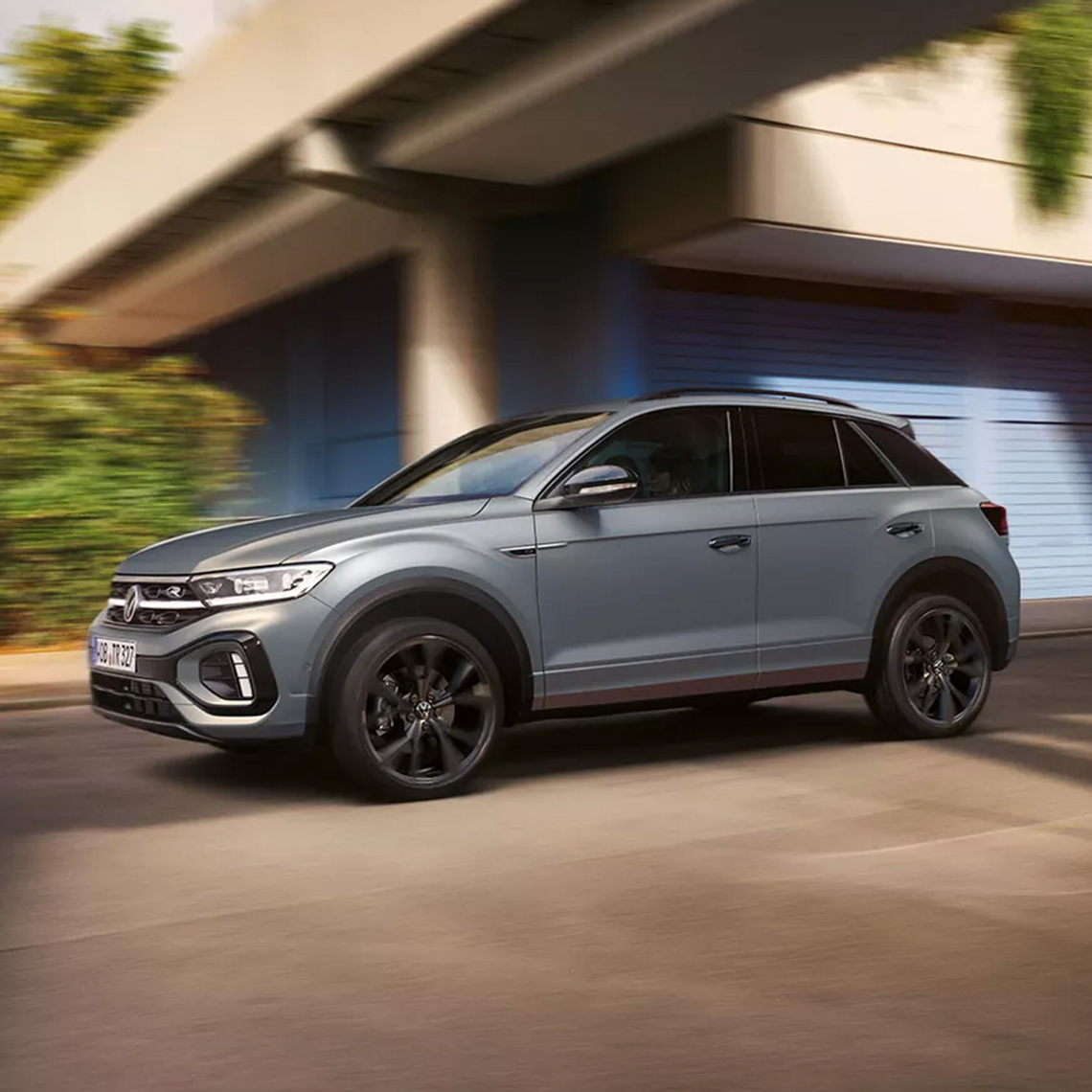 Grauer VW T-Roc R-Line in Seitenansicht fahrend auf einer städtischen Straße vor moderner Architektur mit blau-grauen Rolltoren.